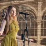 Usmievavá mladá žena v zelených šatách spieva do mikrofónu na pódiu a vrúcne gestikuluje, zatiaľ čo slnečné svetlo vrhá za ňu vzorované tiene.