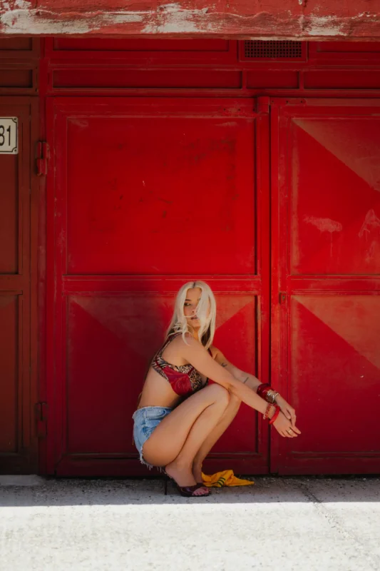 Žena s dlhými blond vlasmi si drepuje pri žiarivo červenej mestskej stene, oblečená v crop tope a džínsových šortkách. Scéna vyžaruje letnú, bezstarostnú atmosféru.