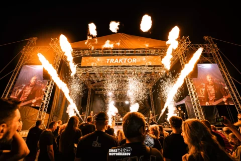 Koncertné pódium v ​​noci s ohnivou pyrotechnikou a živým davom. Nad ním je zobrazený „Traktor“. Energická atmosféra a veľké videoobrazovky ukazujú členov kapely.