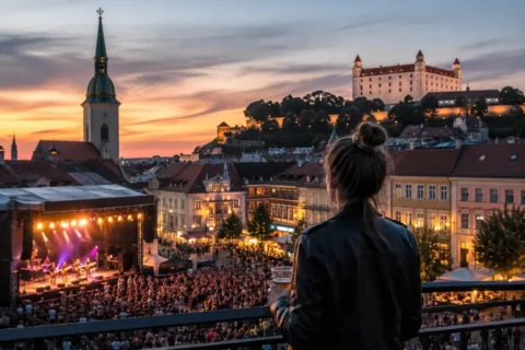 Osoba v čiernej bunde sleduje živý koncert pod holým nebom v Bratislave pri západe slnka, s osvetlenou Katedrálou svätého Martina a Bratislavským hradom.