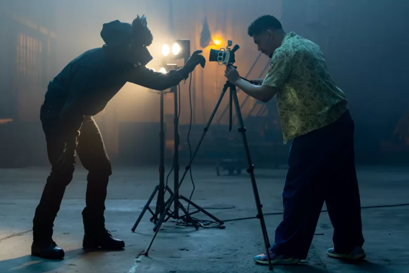 Dvaja filmári umiestňujú kameru na statív v prítmí štúdia, obklopení jasnými svetlami, čo vytvára sústredenú atmosféru spoločnej tvorby.