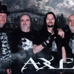 Štyria muži v čiernom rockovom oblečení pózujú pred priemyselnou panorámou zahalenou do dymu; táto atmosférická fotografia metalovej kapely nesie v spodnej časti logo „AXLES“.
