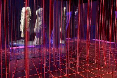 Pohlcujúca umelecká inštalácia žiariacich červených mriežkových čiar a visiacich vertikálnych šnúr obklopuje hmlisté postavy a vytvára surrealistickú, futuristickú atmosféru.