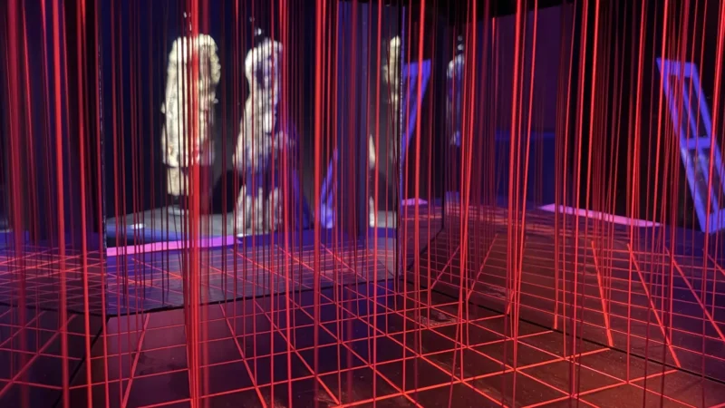 Pohlcujúca umelecká inštalácia žiariacich červených mriežkových čiar a visiacich vertikálnych šnúr obklopuje hmlisté postavy a vytvára surrealistickú, futuristickú atmosféru.