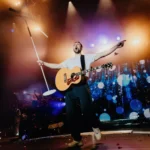 Pesničkár vystupuje na pódiu s akustickou gitarou a stojanom na mikrofón; má rozpažené ruky, zatiaľ čo za ním žiaria farebné svetlá a efekt bokeh.