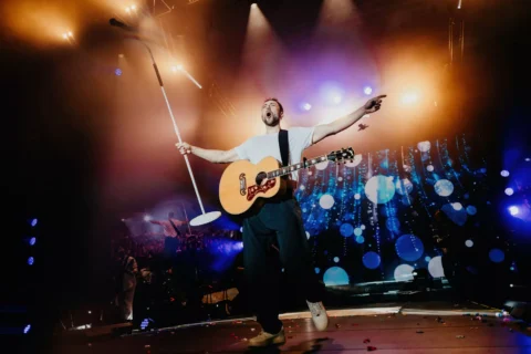 Pesničkár vystupuje na pódiu s akustickou gitarou a stojanom na mikrofón; má rozpažené ruky, zatiaľ čo za ním žiaria farebné svetlá a efekt bokeh.