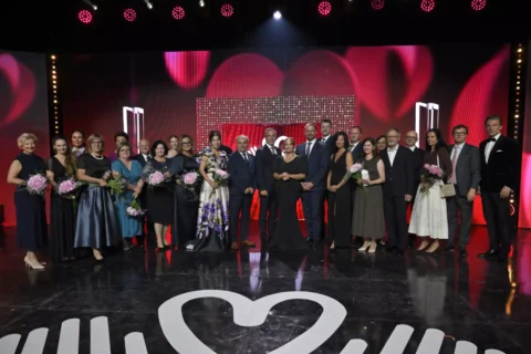 Veľká skupina vo formálnom oblečení pózuje na slávnostnom pódiu a drží kytice pred červeným pozadím s tematikou srdca; slávnostná, elegantná atmosféra.