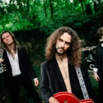 Traja hudobníci v čiernych oblekoch pózujú v bujnom lese: kučeravý gitarista stojí v strede, po boku dvoch členov kapely, čím vytvárajú náladový rockový portrét.