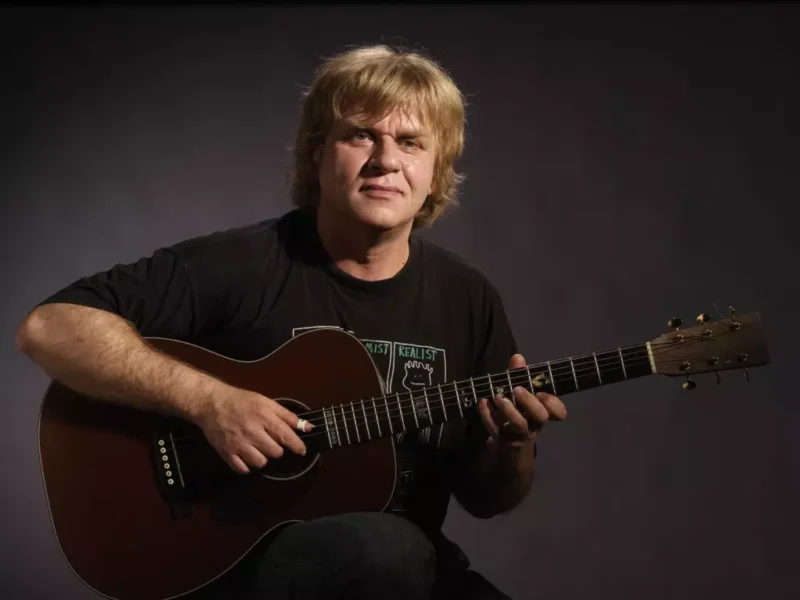 Blondýnsky muž v čiernej košeli hrá na hnedej akustickej gitare na tmavom pozadí s pokojným a sústredeným výrazom.