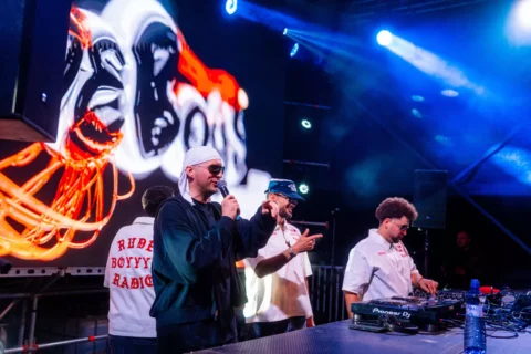 Hip-hopoví interpreti rapujú na pódiu po boku DJ-a; scénu dotvárajú modré svetlá, grafika v štýle graffiti a živá, energická koncertná atmosféra.