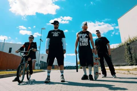 Štyria priatelia pózujú na slnečnej mestskej ulici: jeden s BMX bicyklom, jeden drží skateboard a dvaja stoja neďaleko a vyžarujú sebavedomie.