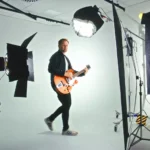 Záber zo zákulisia štúdia zachytávajúci usmievavého gitaristu v čiernom, ktorý hrá na oranžovej elektrickej gitare a je obklopený svetlami, stojanmi a čiernymi pozadiami.