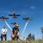 Rocková kapela vystupuje na trávnatej lúke pod štyrmi akrobatickými lietadlami, ktoré za sebou zanechávajú stopy bieleho dymu; nechýbajú gitary, bicie, modrá obloha a energická atmosféra.