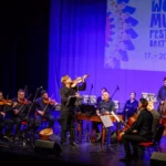 Huslista vedie sláčikový súbor s kontrabasom pod transparentom festivalu World Music; fialové osvetlenie vytvára živú koncertnú atmosféru.