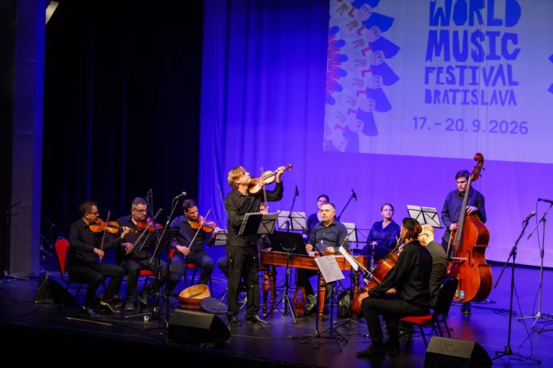 Huslista vedie sláčikový súbor s kontrabasom pod transparentom festivalu World Music; fialové osvetlenie vytvára živú koncertnú atmosféru.