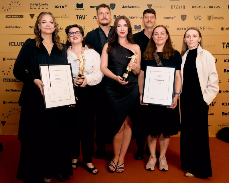 Sedem ocenených pózuje na červenom koberci s trofejami a certifikátmi v rámoch; usmievajú sa pred zlatým pozadím podujatia s logami sponzorov.