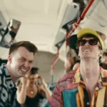 Veselí priatelia sa smejú a tancujú v mestskom autobuse s červenými zábradliami; muž v slnečných okuliaroch a žltom klobúku vedie živú skupinu.