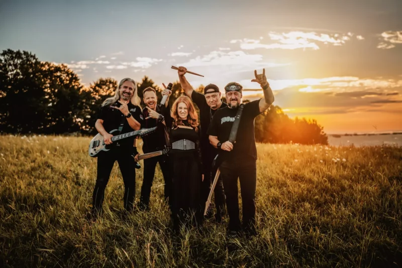 Veselá rocková kapela pózuje s gitarami na slnkom zaliatom poli pri západe slnka a dvíha ruky, zatiaľ čo zlatisté svetlo vytvára hrejivú, slávnostnú atmosféru.
