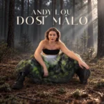 Žena v čiernom tope a splývavej zelenej sukni sedí medzi vysokými lesnými stromami; slnečné lúče a názov „Andy Lou: Dost málo“ vytvárajú dramatickú atmosféru.