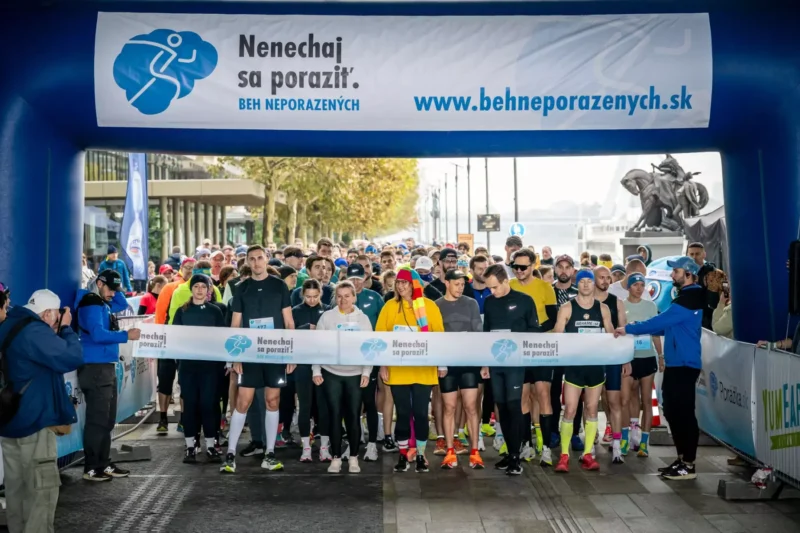 Pod modrou štartovou bránou sa zhromažďuje dav bežcov, zatiaľ čo dobrovoľníci držia štartovaciu pásku, čím na komunitnom bežeckom podujatí vytvárajú optimistickú a odhodlanú atmosféru.