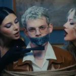Zviazaný blond muž sedí medzi dvoma ženami, ktoré sa k sebe nakláňajú; ústa mu zakrýva páska s nápisom „CHEATER“ (PODRÁDAČ), zatiaľ čo jedna z nich ukazuje prstom a vytvára tak napätú, inscenovanú atmosféru.