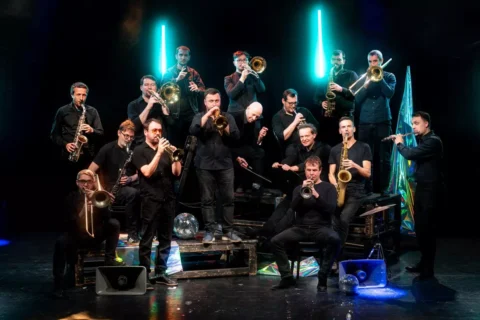 Na tmavom pódiu vystupuje energický džezový súbor; hudobníci hrajú na saxofóny, trúbky a trombón pod žiarivými tyrkysovými svetlami.