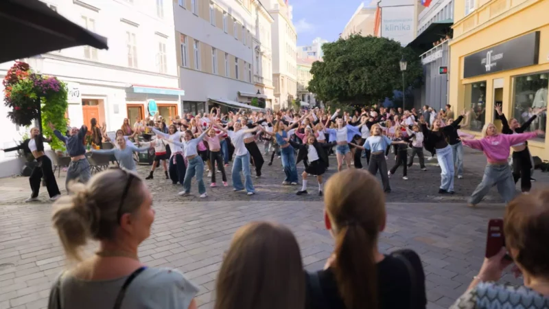 Živé tanečné podujatie pod holým nebom zapĺňa pešiu zónu; dav napodobňuje pohyby inštruktorov, zatiaľ čo v popredí všetko sledujú diváci.
