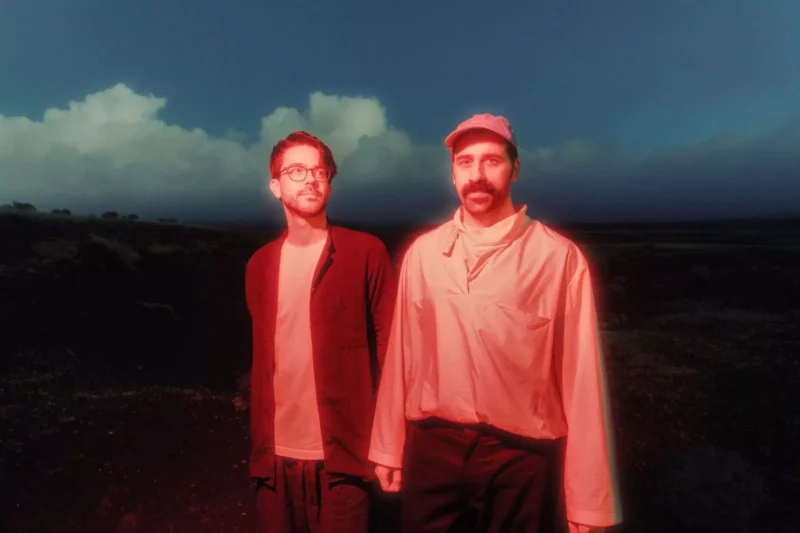 Two men stand in a dark, open landscape beneath cloudy skies, lit in vivid red; their still poses create an eerie, cinematic mood.