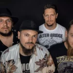 Štyria členovia Alter Bridge pózujú na čiernom pozadí v tmavých tričkách a klobúkoch; ich výrazy vytvárajú vážnu a sebavedomú rockovú atmosféru.