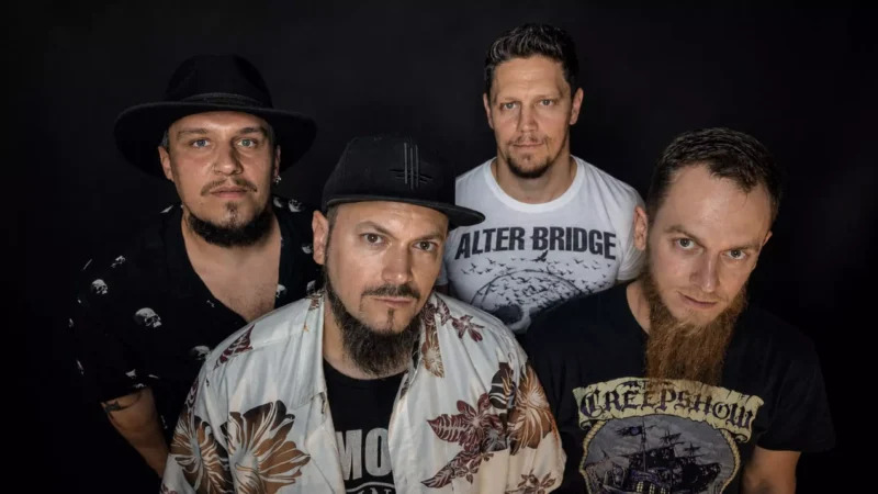 Štyria členovia Alter Bridge pózujú na čiernom pozadí v tmavých tričkách a klobúkoch; ich výrazy vytvárajú vážnu a sebavedomú rockovú atmosféru.