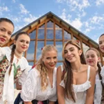 Šesť usmievavých mladých žien vo farebných ľudových krojoch a bielych blúzkach pózuje pred drevenou stavbou pod jasnou modrou oblohou, pričom vyžarujú teplo a slávnostnú náladu.