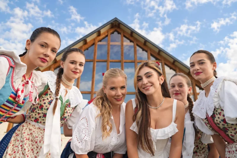 Šesť usmievavých mladých žien vo farebných ľudových krojoch a bielych blúzkach pózuje pred drevenou stavbou pod jasnou modrou oblohou, pričom vyžarujú teplo a slávnostnú náladu.