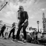 Čiernobiely pouličný koncert: speváčka sa nakláňa k mikrofónu, zatiaľ čo gitarista a bubeník hrajú na mestskom námestí, čím vytvárajú energickú, a pritom komornú atmosféru.