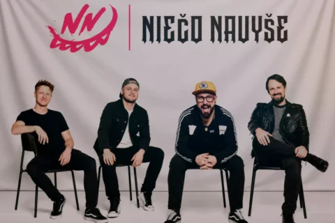 Štyria muži sedia na stoličkách pred svetlým pozadím, pod červeným logom a názvom „Ničo Novuše“; pózujú ležérne v čiernom oblečení a pôsobia ako uvoľnená hudobná skupina.