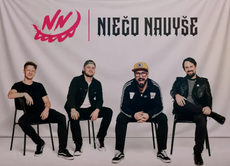 Štyria muži sedia na stoličkách pred svetlým pozadím, pod červeným logom a názvom „Ničo Novuše“; pózujú ležérne v čiernom oblečení a pôsobia ako uvoľnená hudobná skupina.