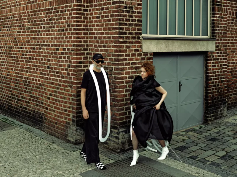 Hravý módny editoriál zachytáva dve modelky v čiernych outfitoch pri tehlovom múre a kovových dverách; do popredia vystupujú dlhý biely šál a biele čižmy.