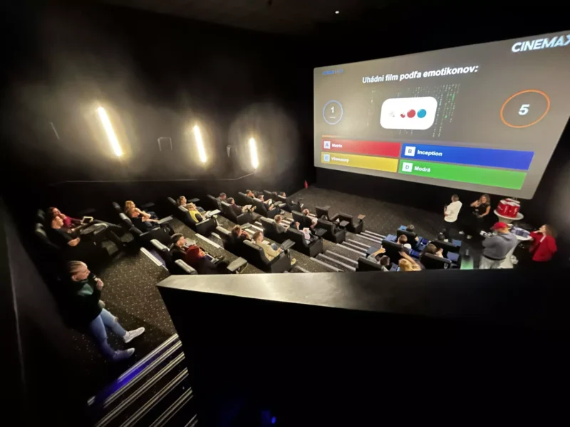 Moderná kinosála s divákmi sediacimi v radoch, obrátenými k veľkému plátnu, na ktorom sa premieta farebný filmový kvíz; atmosféra pôsobí živo a interaktívne.