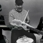 Čiernobiela fotografia troch vystupujúcich hudobníkov: basgitaristu v obleku, bubeníka v pruhovanej košeli a gitaristu so slnečnými okuliarmi. Ich energické pózy vytvárajú živú atmosféru rockovej kapely.