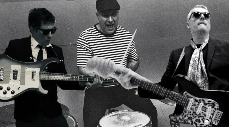 Čiernobiela fotografia troch vystupujúcich hudobníkov: basgitaristu v obleku, bubeníka v pruhovanej košeli a gitaristu so slnečnými okuliarmi. Ich energické pózy vytvárajú živú atmosféru rockovej kapely.