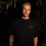 Pokojný, hĺbavý muž v čiernom tričku stojí uprostred tienistého lístia; na tomto dramatickom portréte so slabým osvetlením je vidieť jeho potetované predlaktie.