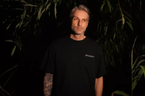 Pokojný, hĺbavý muž v čiernom tričku stojí uprostred tienistého lístia; na tomto dramatickom portréte so slabým osvetlením je vidieť jeho potetované predlaktie.