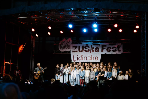 Na festivale Zuška Fest sa na pódiu pod červenými a modrými svetlami zhromažďuje veľký zbor; naľavo stojí gitarista a v popredí vidno siluety publika – vládne slávnostná atmosféra.